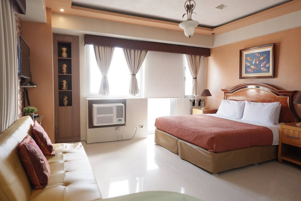 Une chambre d’hôtel comprenant un lit, un canapé, une table de chevet, une lampe, une fenêtre avec des rideaux, un ventilateur de plafond et une étagère murale décorative.