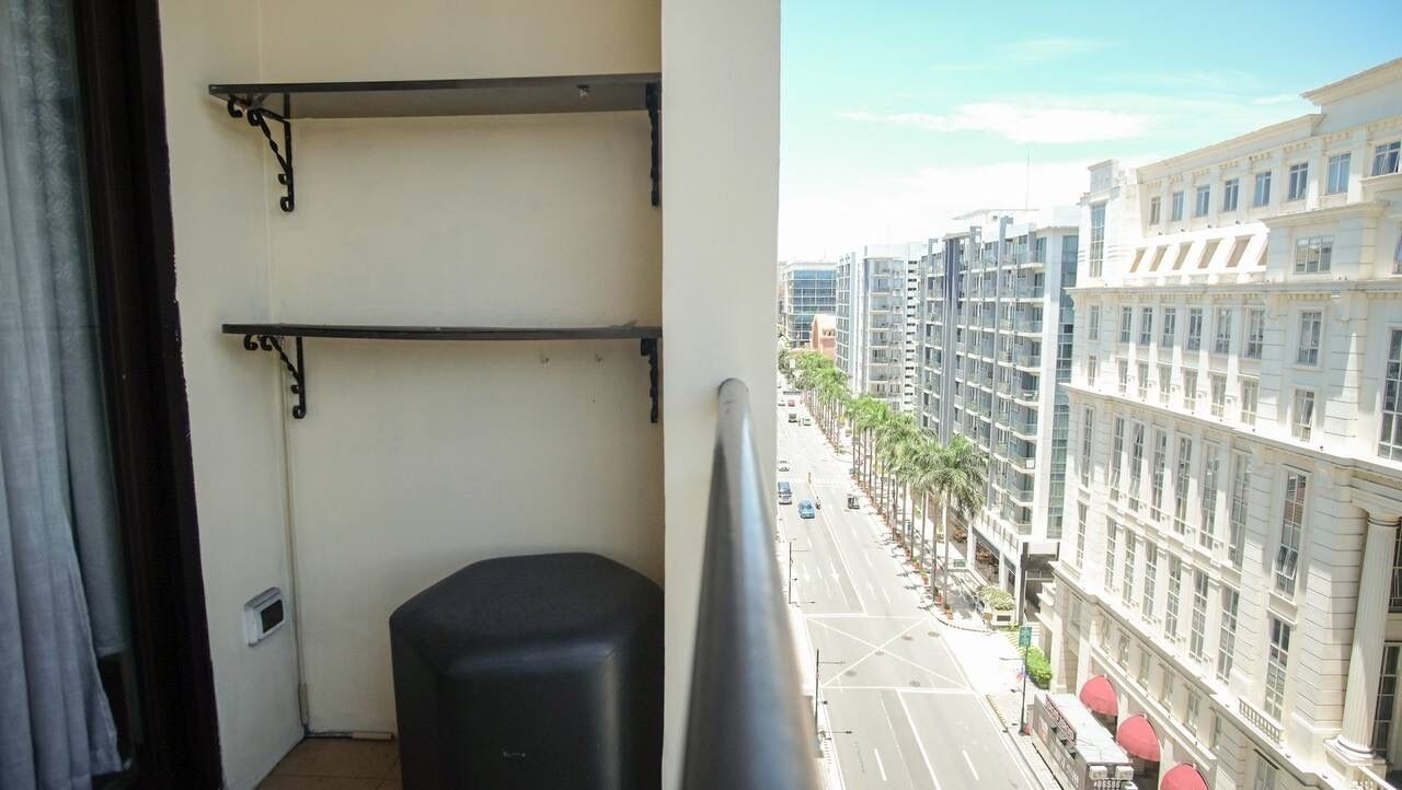 A balcony with metal railings, a black chair, and a view of a city street with buildings and palm trees.