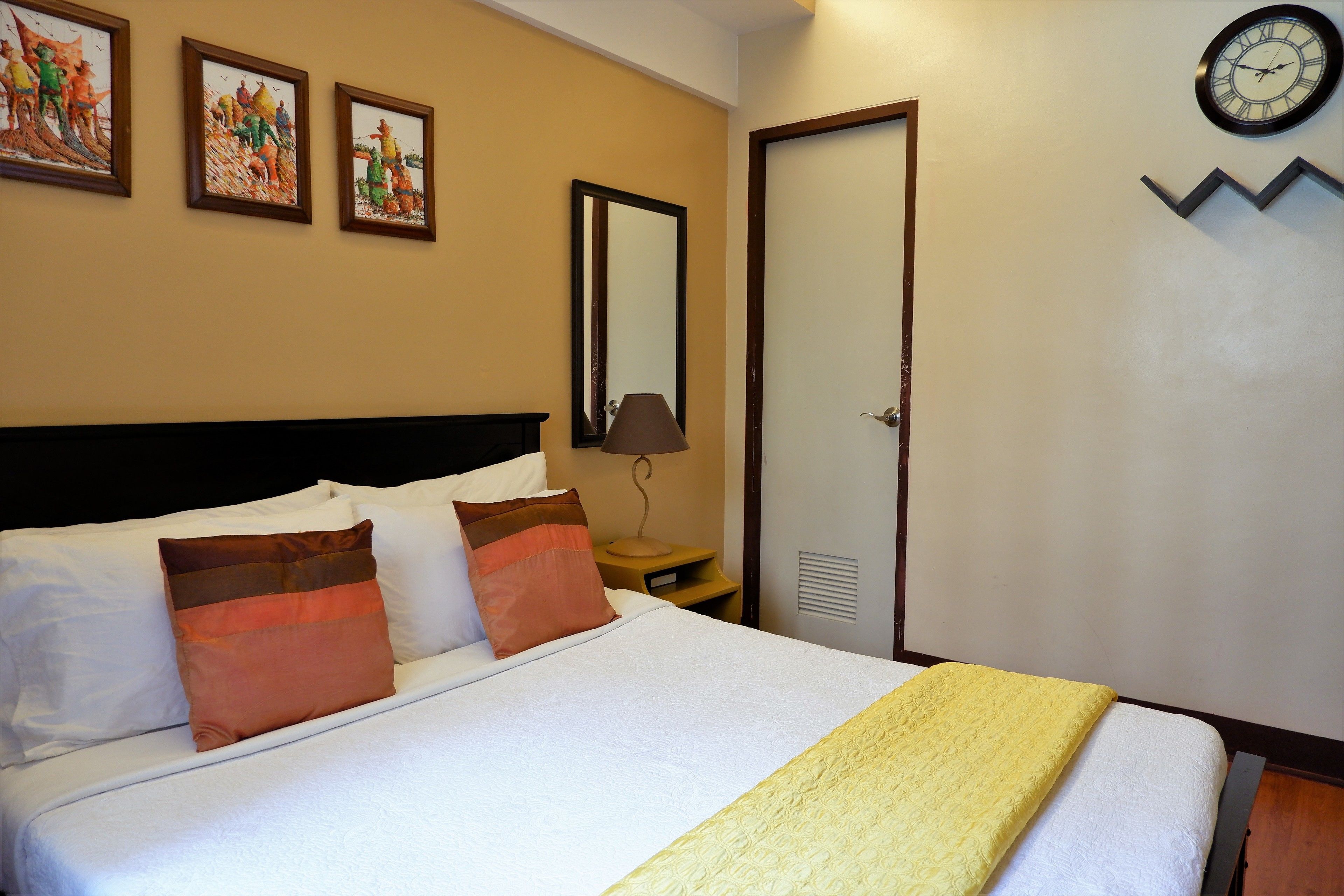 A hotel room with a bed, two pillows, a nightstand, a lamp, a mirror, a clock, and a door.