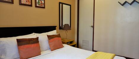 A hotel room with a bed, two pillows, a nightstand, a lamp, a mirror, a clock, and a door.