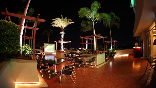 An outdoor dining area with tables and chairs, surrounded by palm trees and plants, under a covered structure with a view of the city at night. - Rathna Residency (Coimbatore)