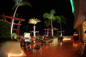 Un área de comedor al aire libre con mesas y sillas, rodeada de palmeras y plantas, bajo una estructura cubierta con vista a la ciudad de noche.