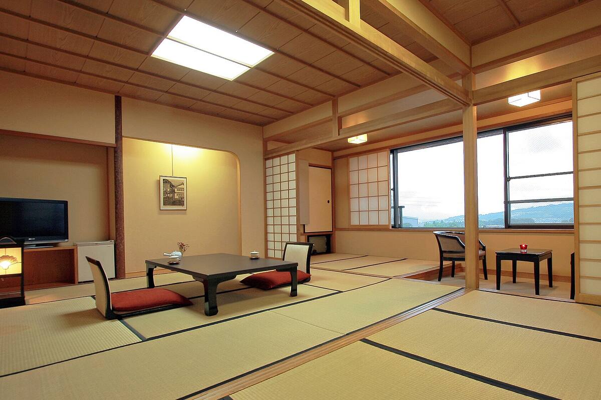 A traditional Japanese room with tatami flooring, a low table with cushions, a television, and a window with a view.