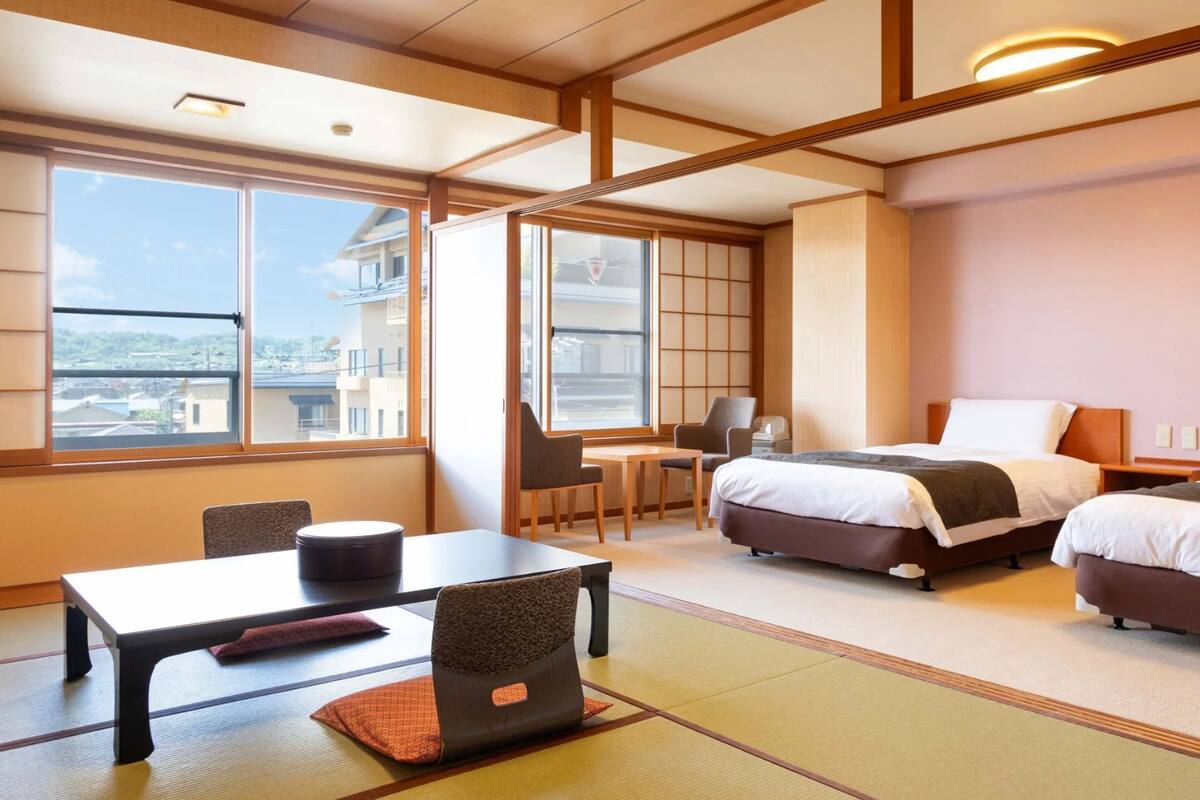 A hotel room with two beds, a desk, a chair, and large windows.