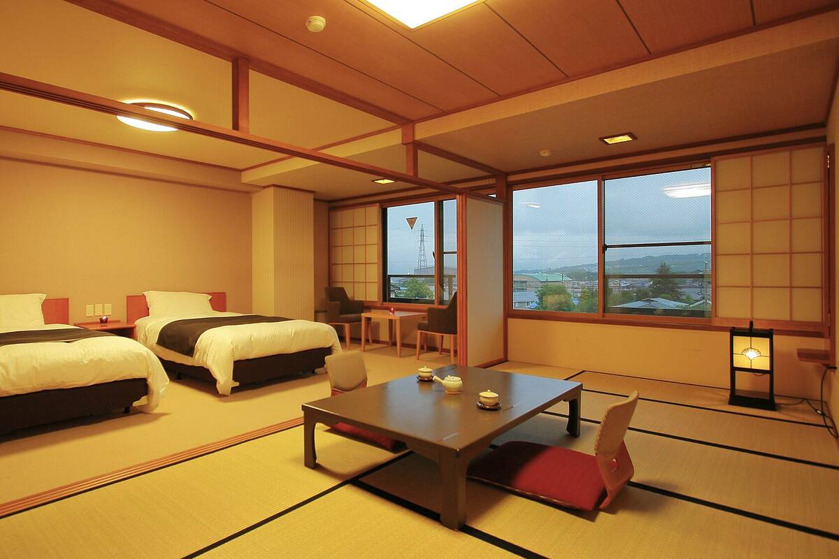 A hotel room with two beds, a small table, a chair, and large windows with a view.