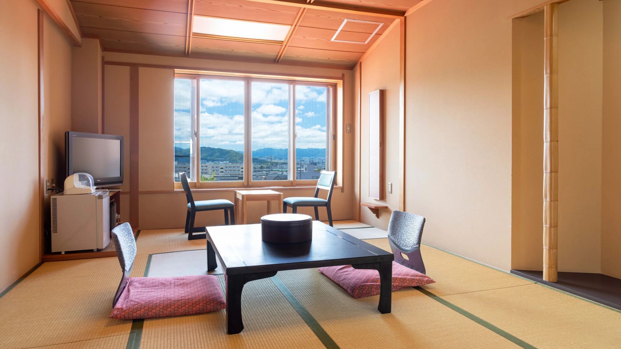 A room with tatami mats, a table, chairs, a television, and a view of a cityscape.