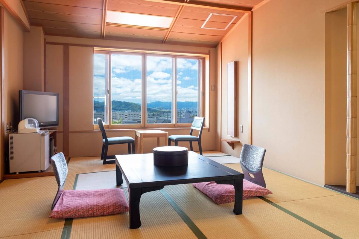 A room with tatami mats, a table, chairs, a television, and a view of a cityscape.