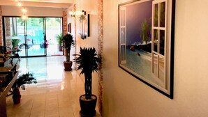 Un hall d’hôtel avec un sol carrelé, des plantes en pot, un tableau encadré représentant une scène de plage et des luminaires fixés au mur.