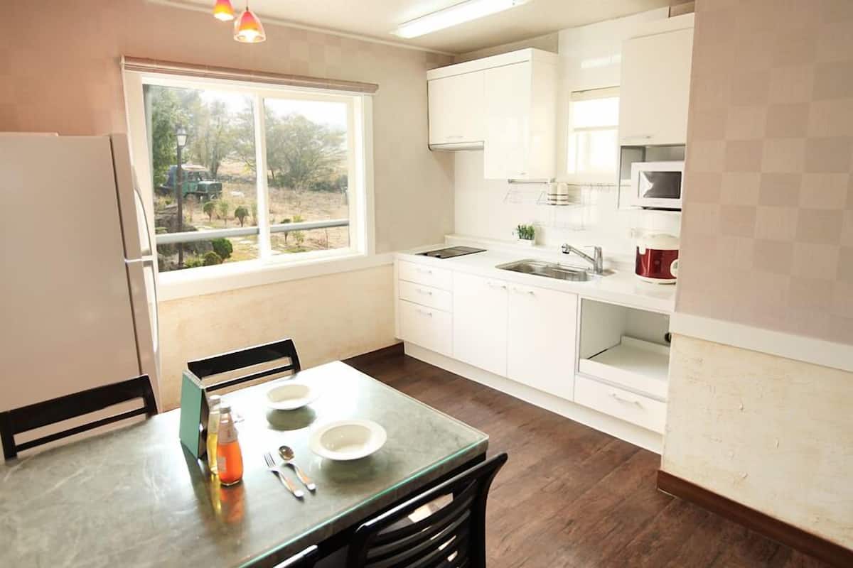 A compact kitchen with white cabinets, a dining table with chairs, a window with a view of greenery, and a refrigerator.