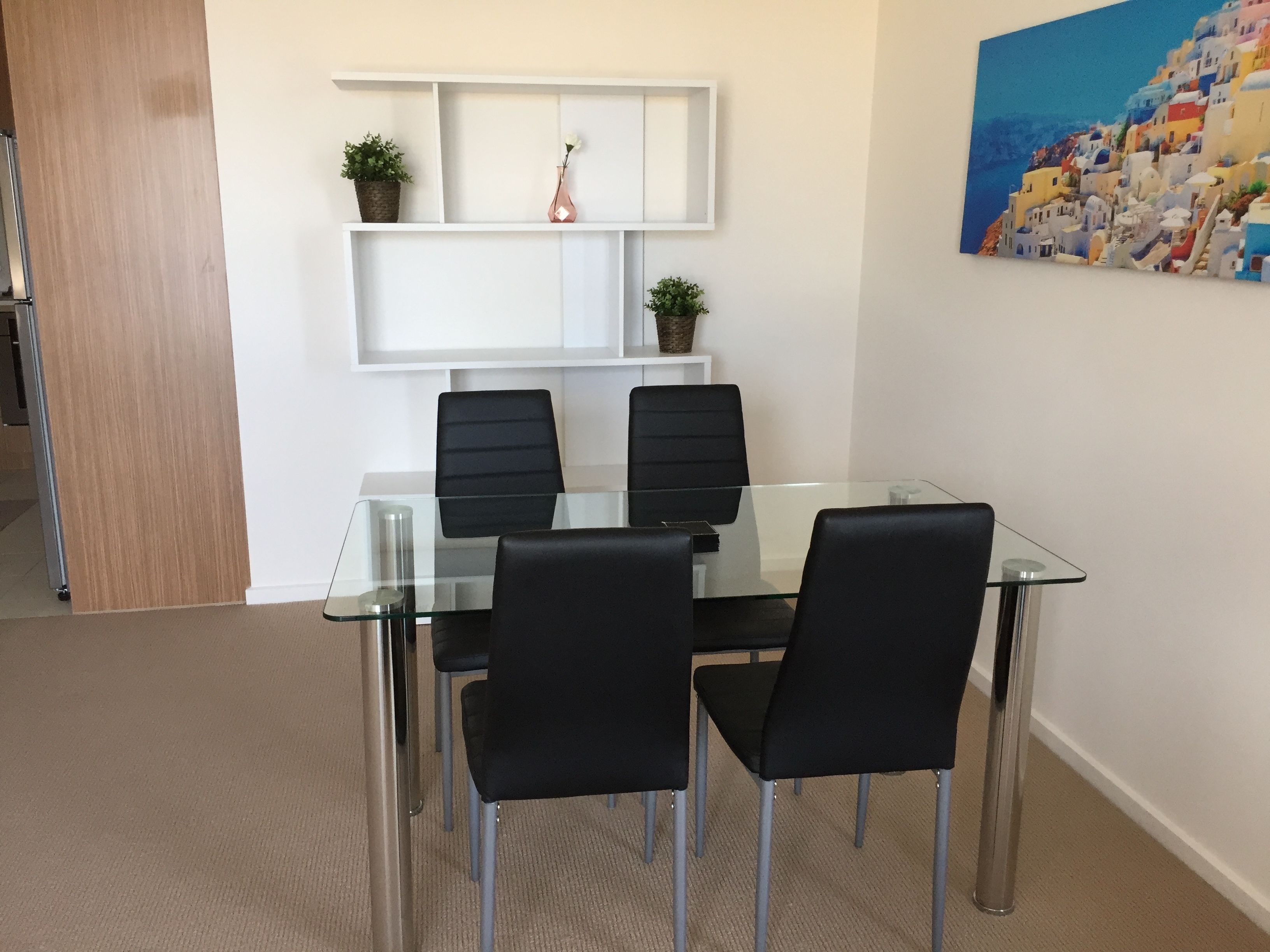A meeting room with a glass table and four black chairs, a white shelf with plants, and a painting of a coastal village on the wall.