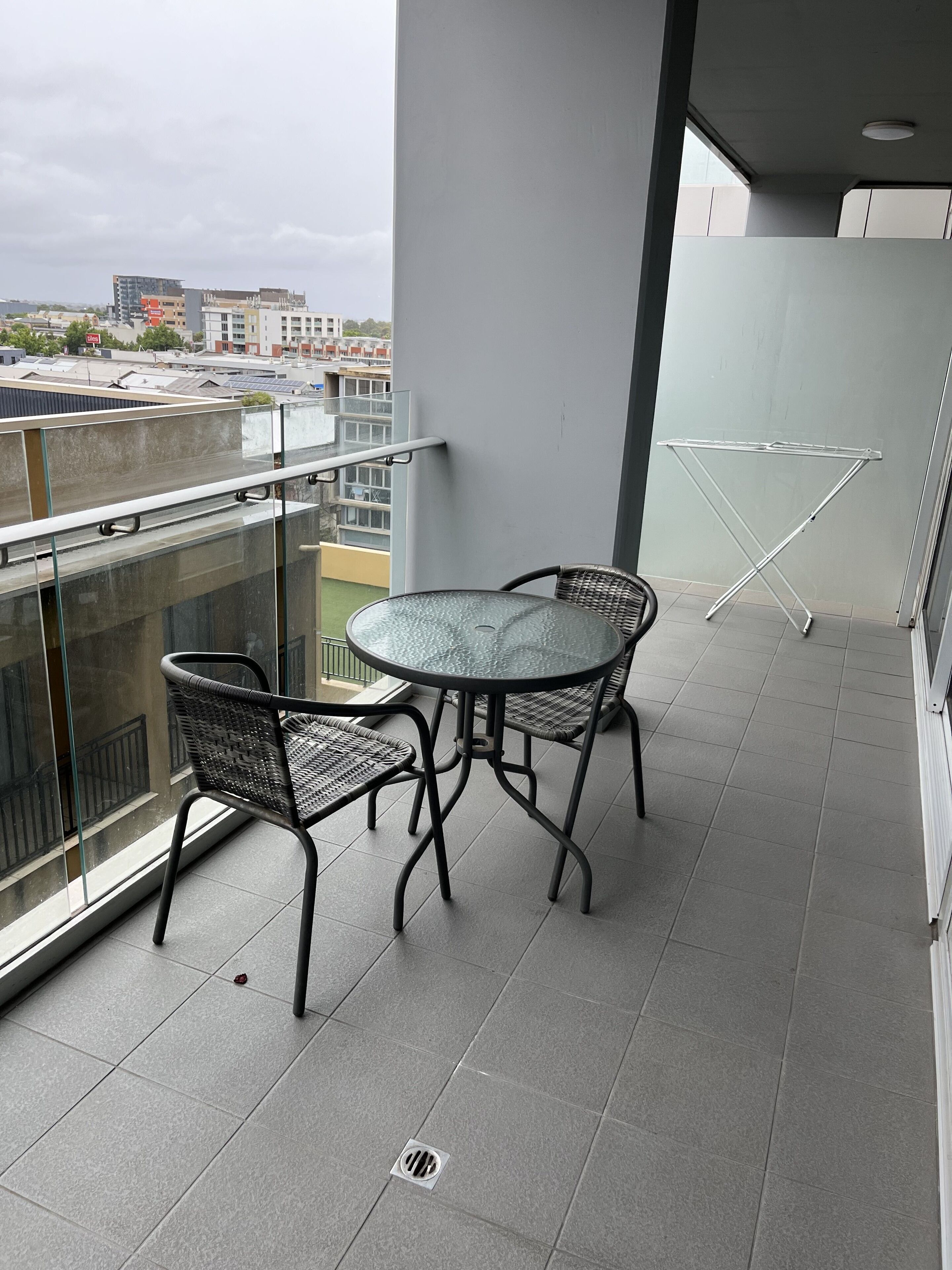 A balcony with a glass table and two wicker chairs, a metal drying rack, and a view of the cityscape.