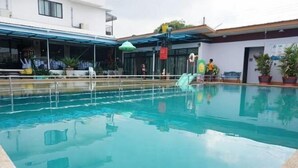 Una piscina con superficie de agua azul cristalina, rodeada por un área techada con plantas en macetas y un edificio al fondo.