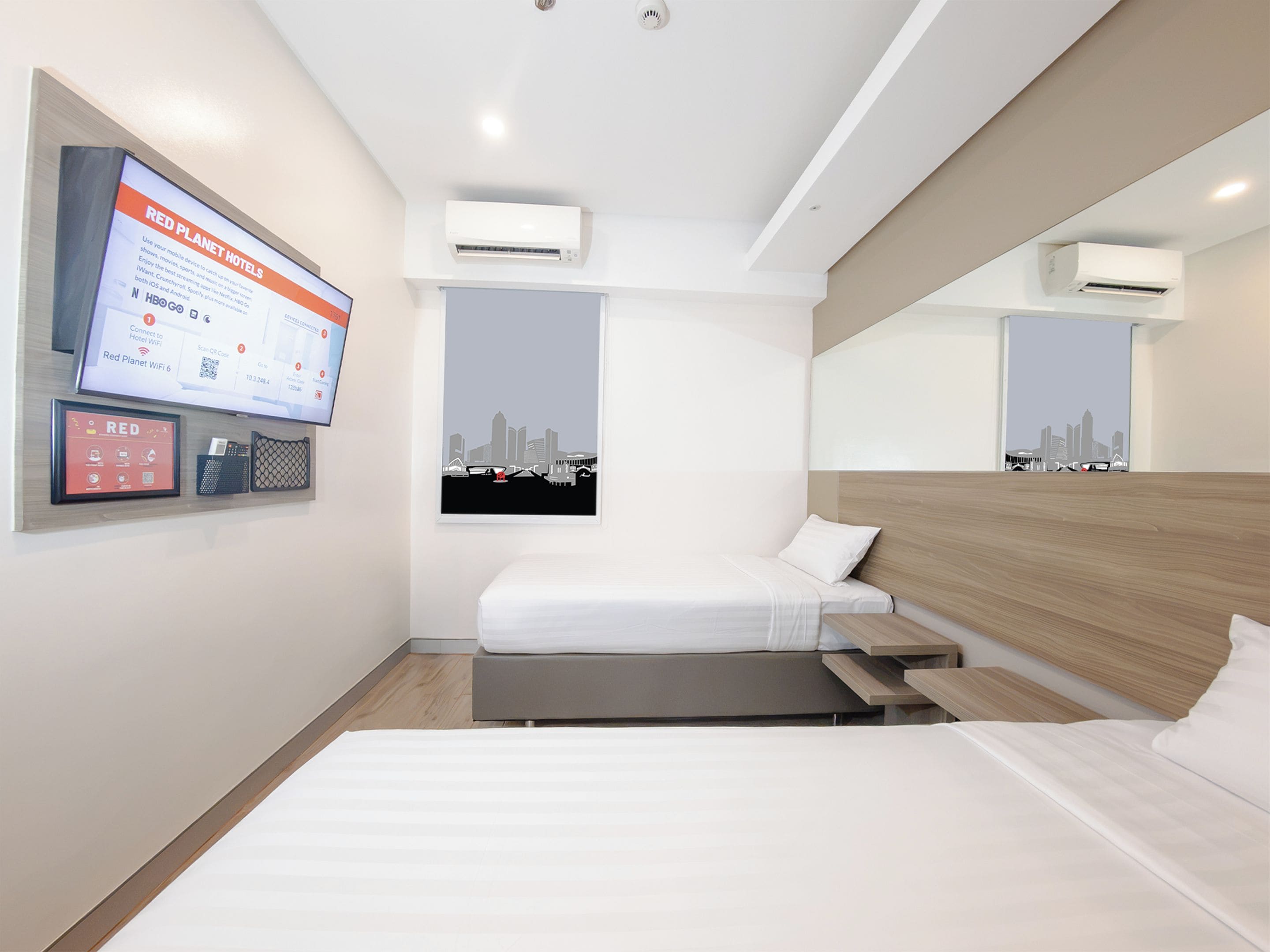 A hotel room with a bed, a desk, a TV mounted on the wall, and two air conditioning units.