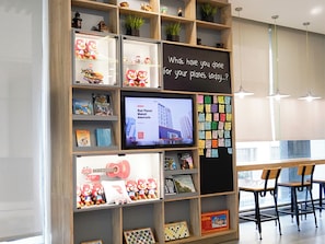 A room with a television displaying a cityscape, a blackboard with a question, shelves with books and toys, and a dining area with chairs.