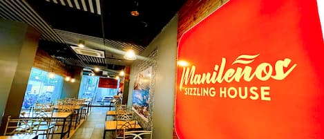 A restaurant interior with tables and chairs, a red wall with the name 'Manilenos Sizzling House,' and a view of the street outside.