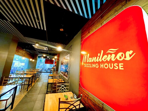 Interior de un restaurante con mesas y sillas, una pared roja con el nombre 'Manilenos Sizzling House' y vista de la calle.
