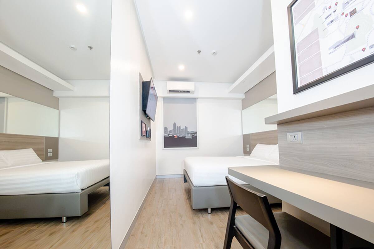 A modern hotel room with two beds, a desk, a chair, and a cityscape poster.