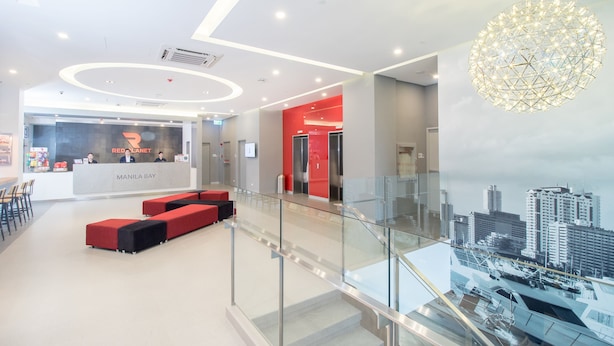 A modern hotel lobby with a reception desk, red seating, and a cityscape view from the upper level.