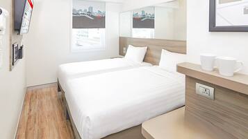 Habitación de hotel con cama, mesita de noche con dos tazas blancas, espejo y ventana con vista a la ciudad.