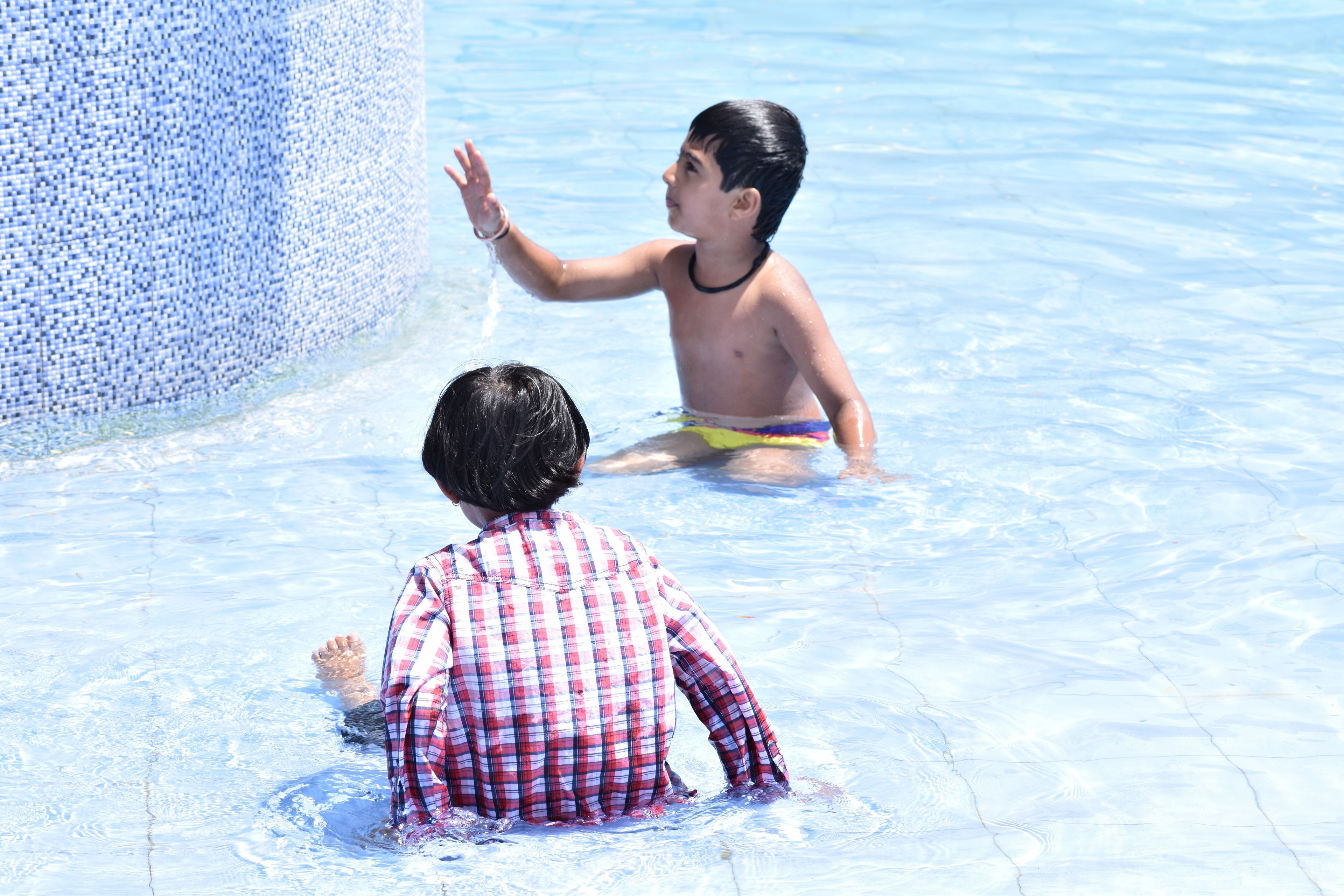 Una piscina con agua azul cristalina, bordeado de azulejos, y un niño con camisa y bañador a cuadros.