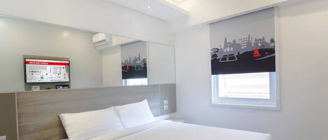A modern hotel room with a large bed, a mounted TV, a window with a cityscape view, and a built-in desk.