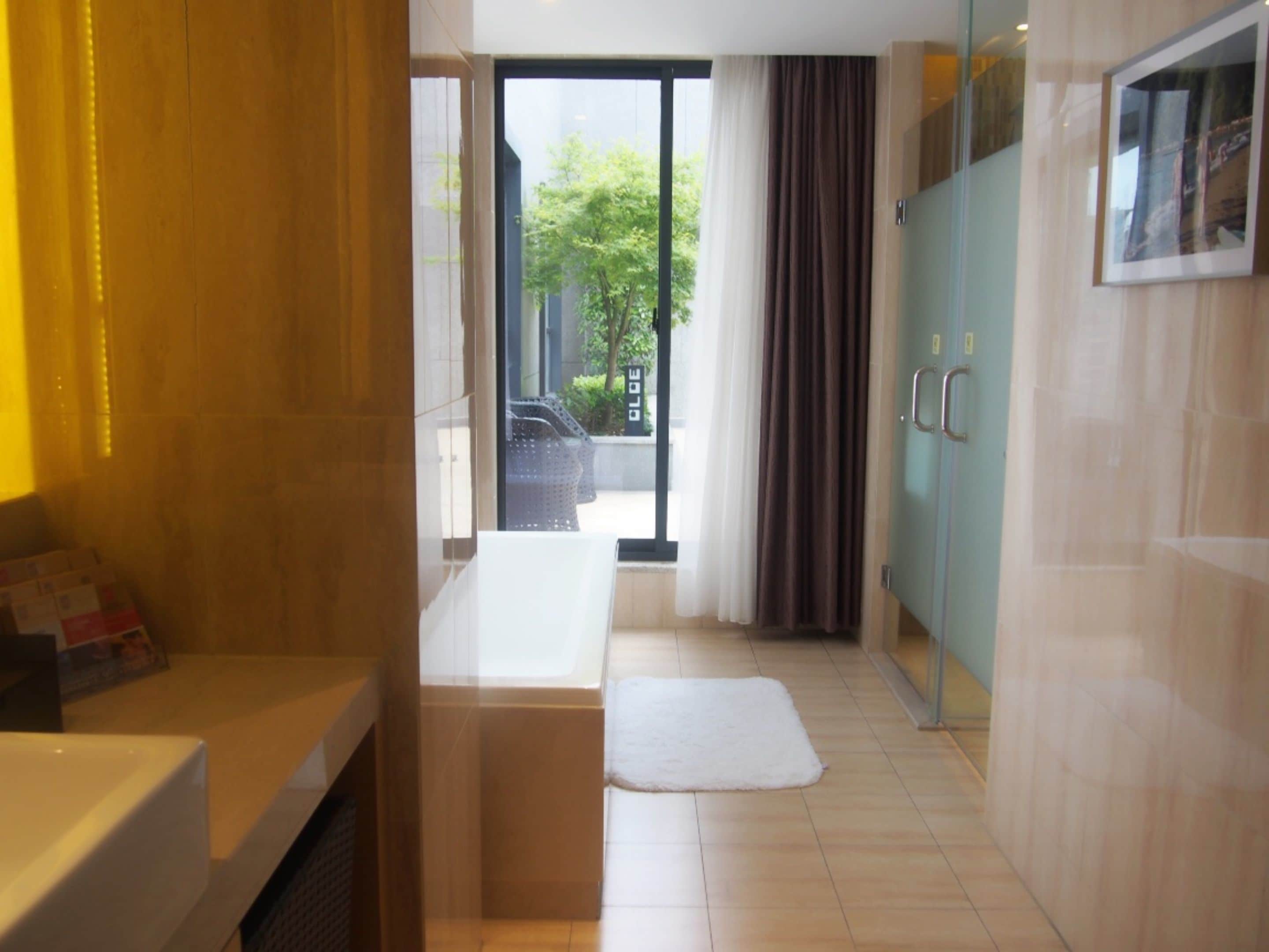 A modern bathroom with a bathtub, a toilet, and a shower area. There is a window with curtains, and a view of the outside.