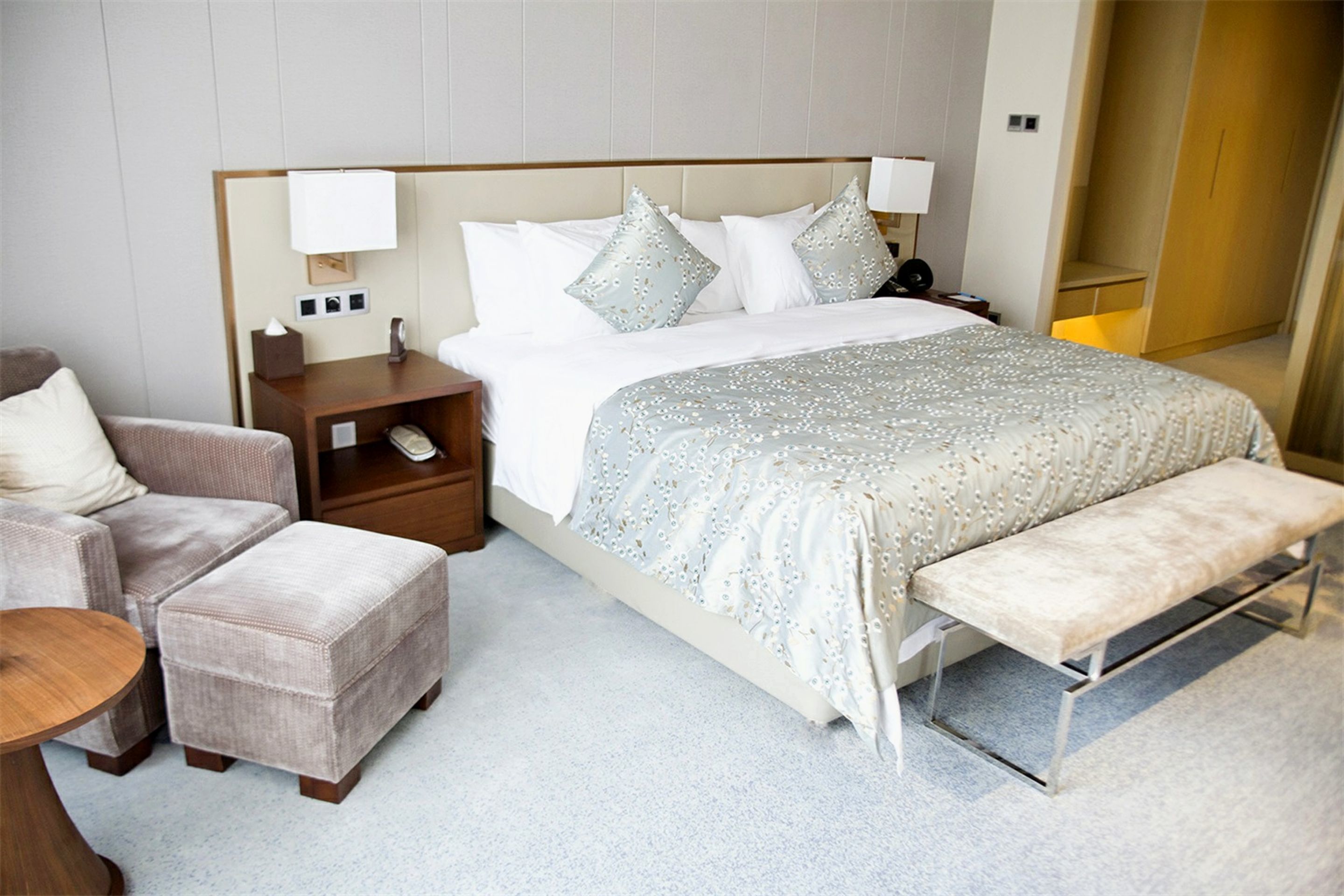 A hotel room with a large bed, a bedside table, a chair, a bench, and a small table.