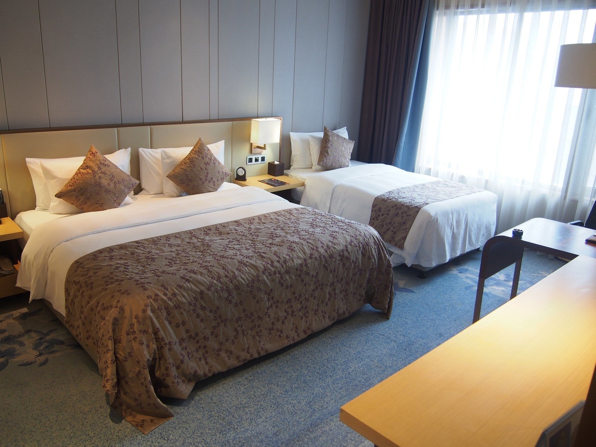 A hotel room with two beds, a desk, and a nightstand with a lamp.