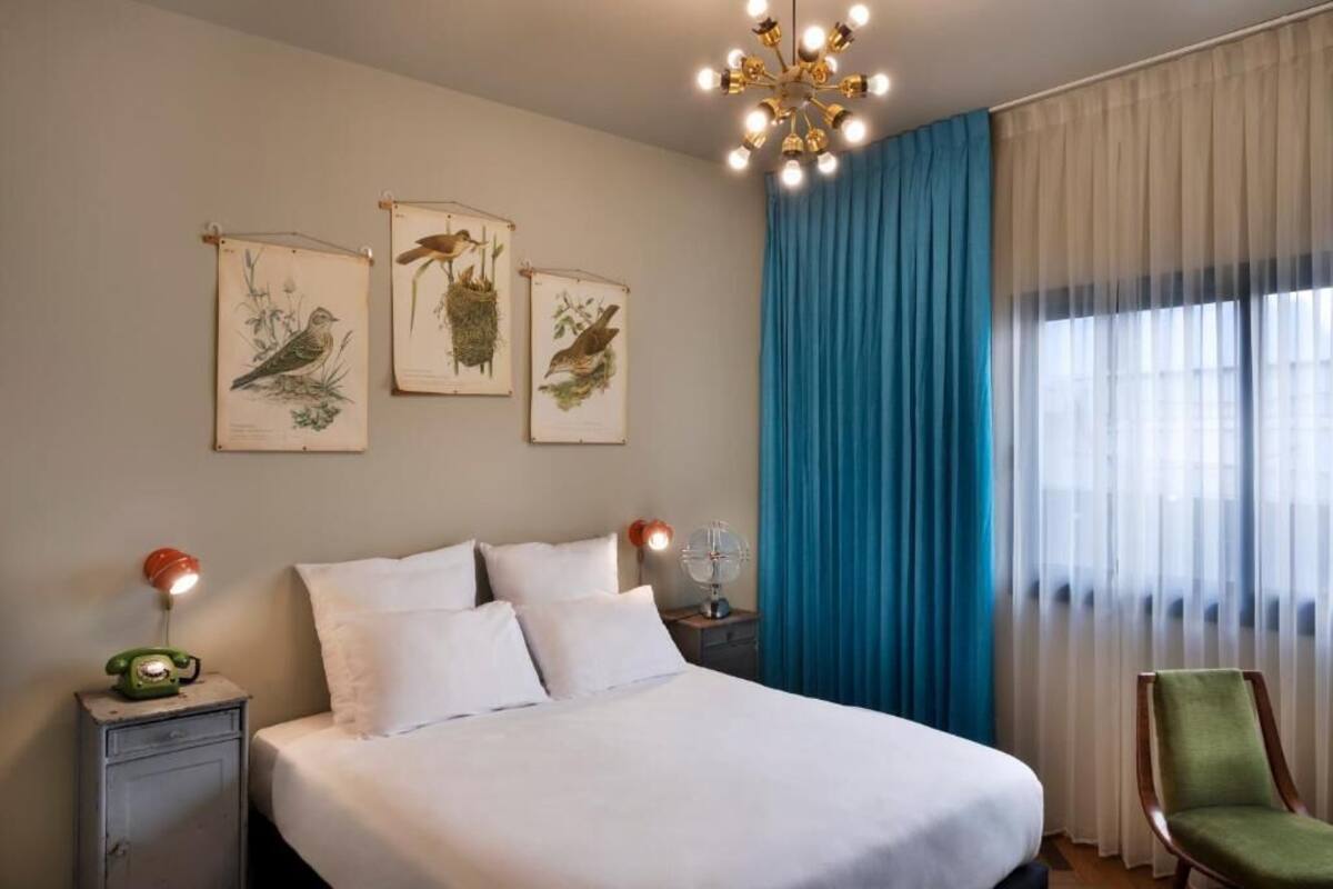 A neatly made bed with white linens, a green chair, a nightstand with a lamp, and wall art depicting birds and plants.
