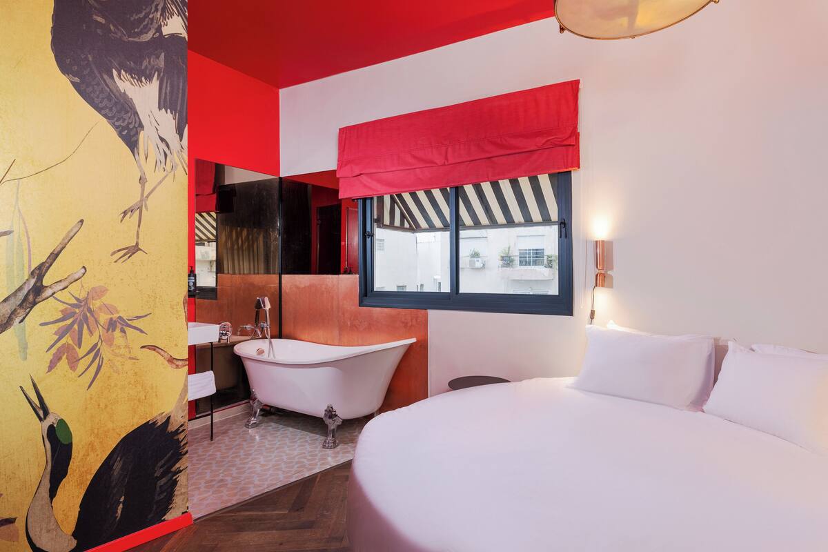 A modern hotel room with a large bed, a bathtub, and a window with a view of the outside.