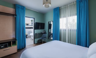 Habitación de hotel con cama, escritorio, silla, televisión y balcón con vistas.