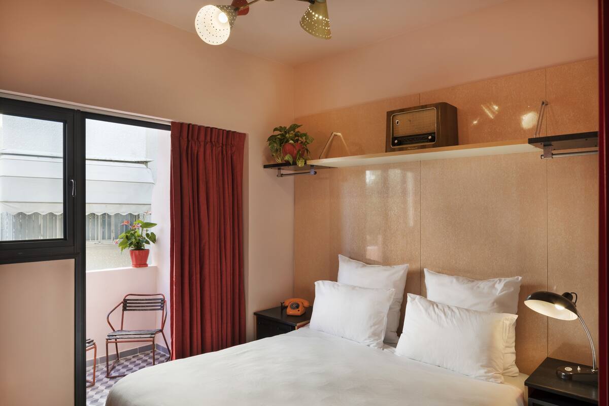 A hotel room with a bed, a nightstand, a radio, a potted plant, a chair, and a window with curtains.