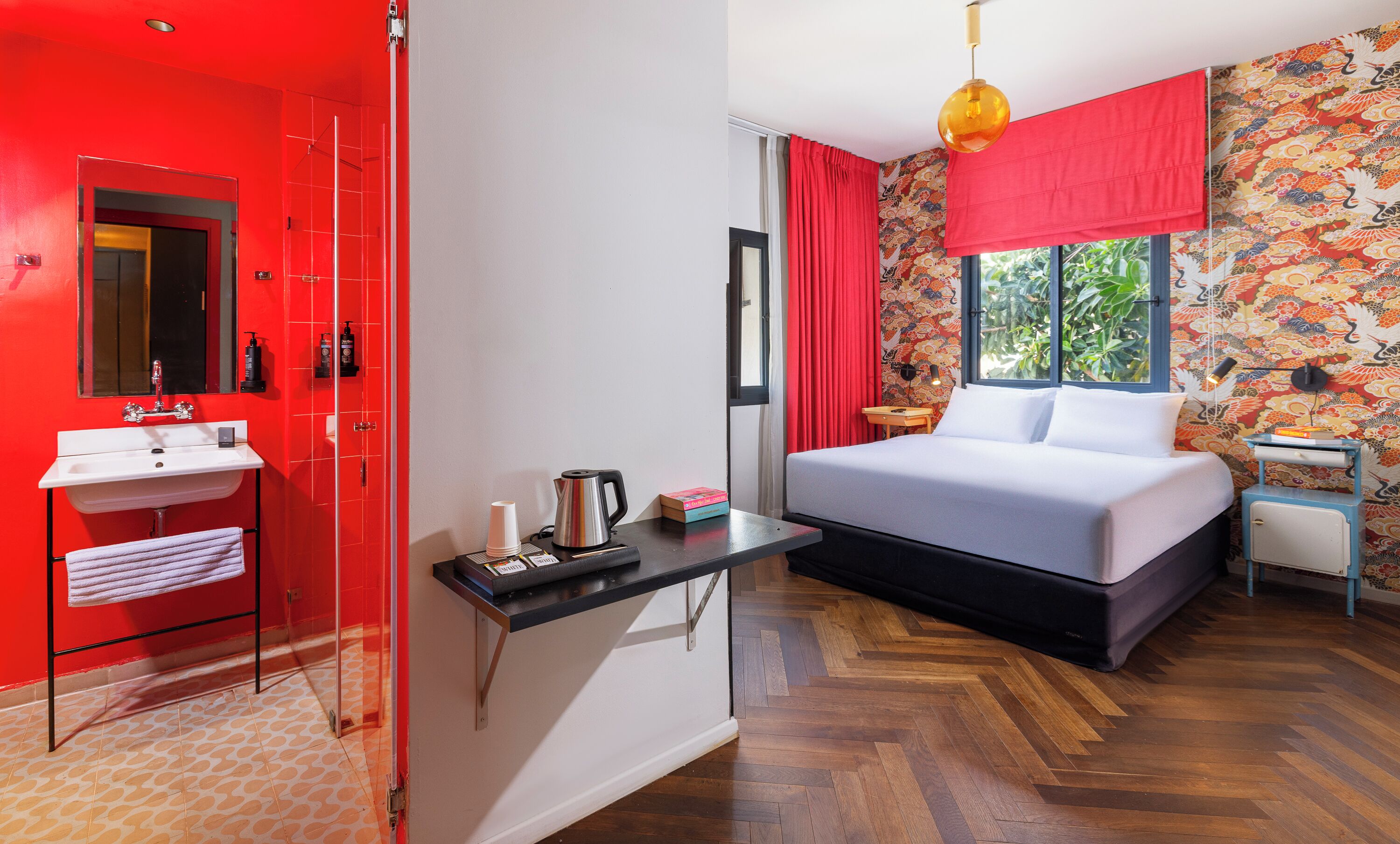 Habitación de hotel con puerta roja, lavamanos blanco, cama con ropa de cama blanca, mesita con hervidor y ventana con cortinas rojas.