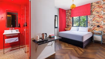 Habitación de hotel con puerta roja, lavamanos blanco, cama con ropa de cama blanca, mesita con hervidor y ventana con cortinas rojas.
