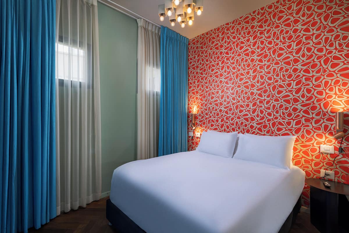 A hotel room with a bed, curtains, a patterned wall, and a nightstand.
