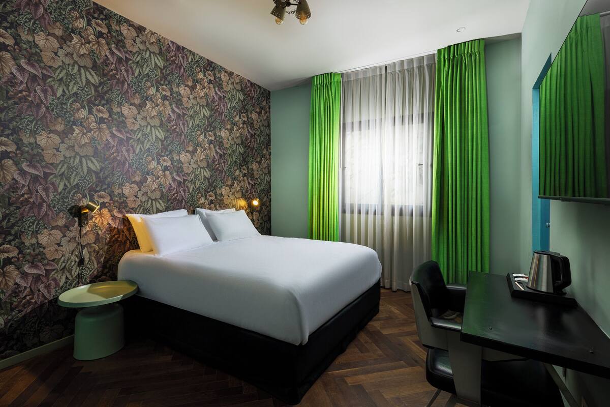 A hotel room with a floral wallpaper, a bed with white linens, green curtains, a desk with a chair, and a small trash bin.