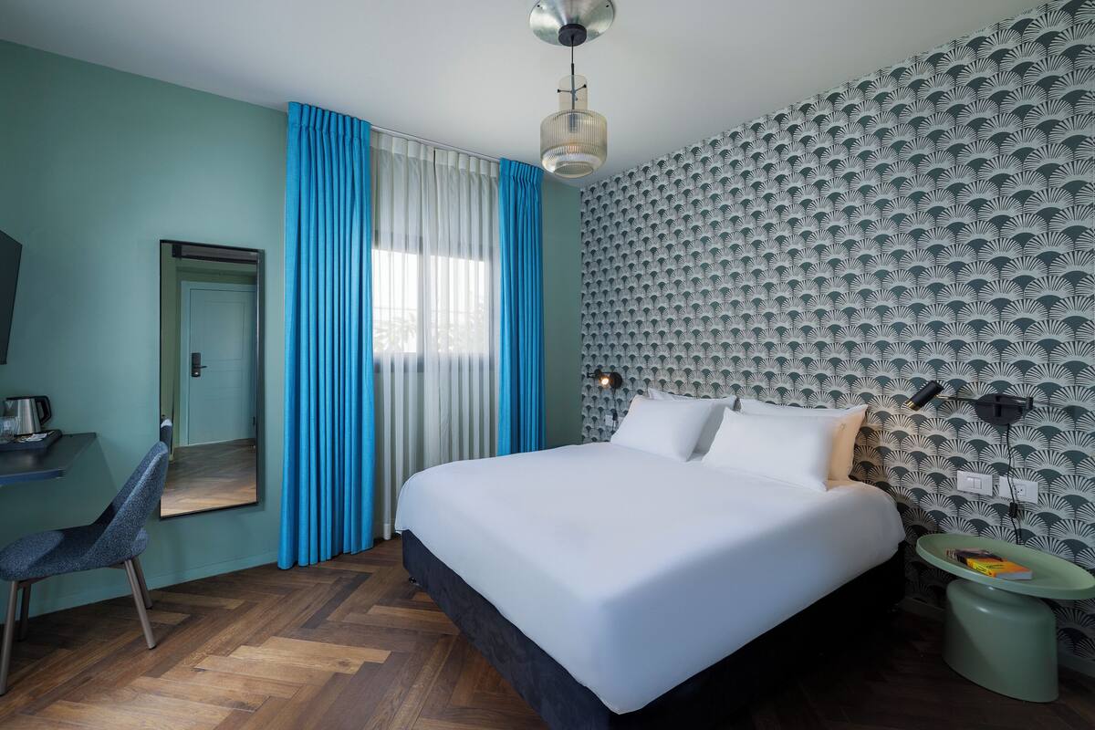 A modern hotel room with a large bed, a desk with a chair, a small table with a lamp, and a wall with a geometric pattern.