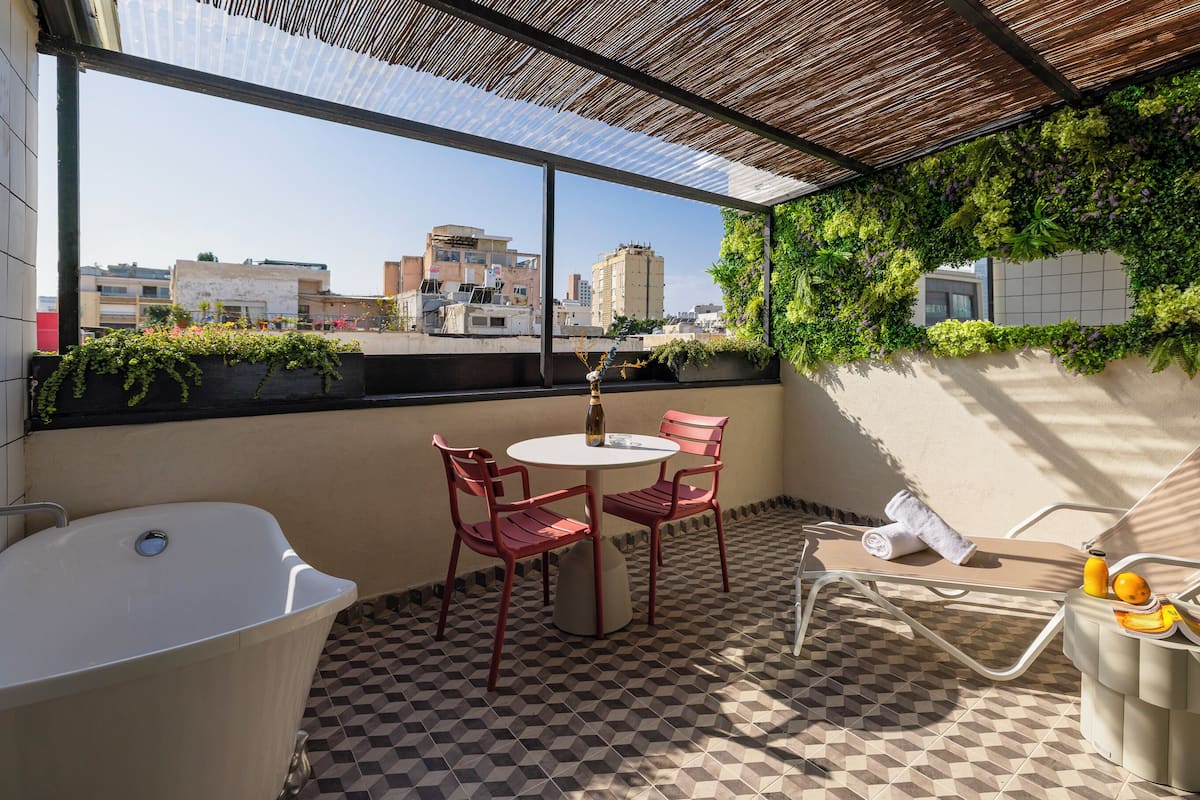 A rooftop terrace with a checkered floor, a white bathtub, two red chairs, a white table, and a glass bottle.