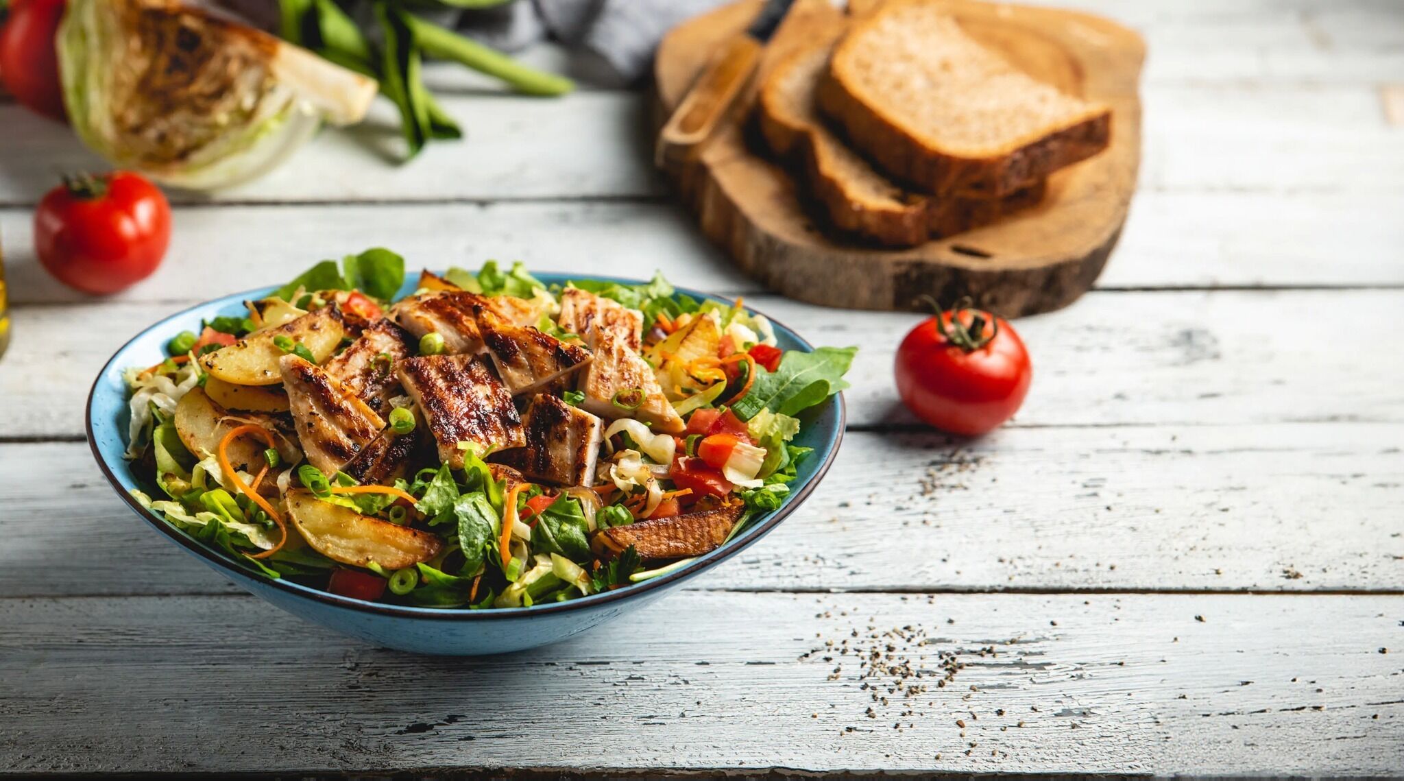 Insalata con pollo alla griglia, verdure e un tagliere di legno con pane sullo sfondo.