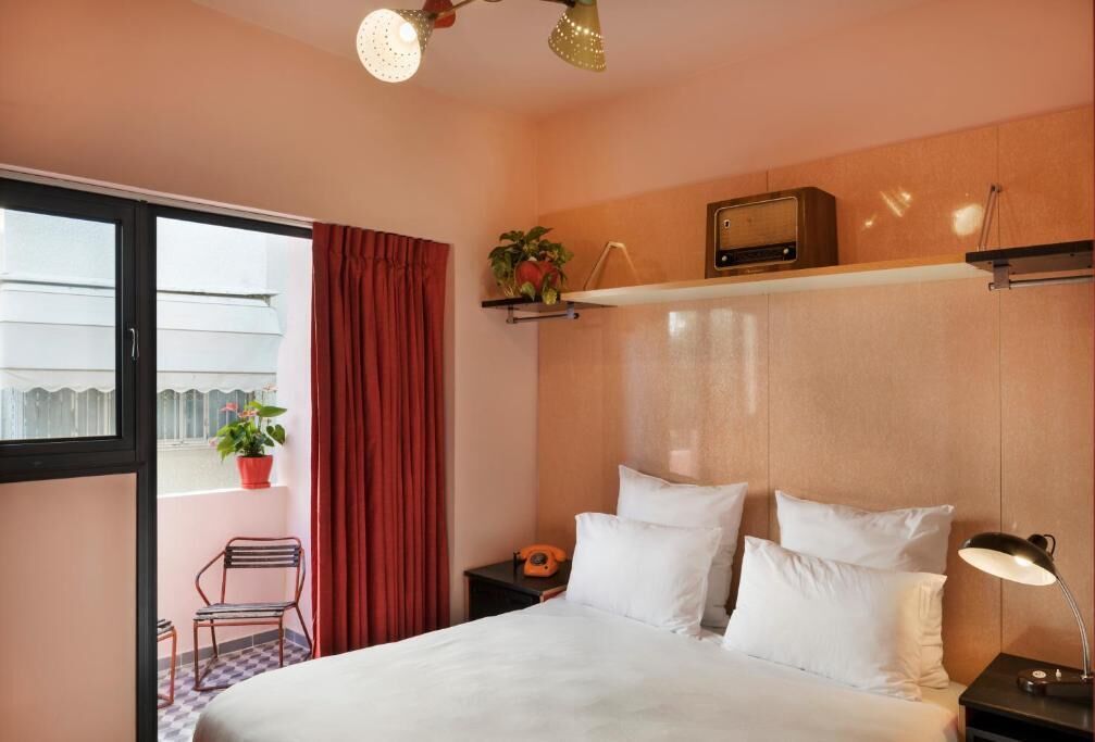 Habitación de hotel con cama, mesita de noche, radio, planta en maceta, silla y ventana con cortinas.