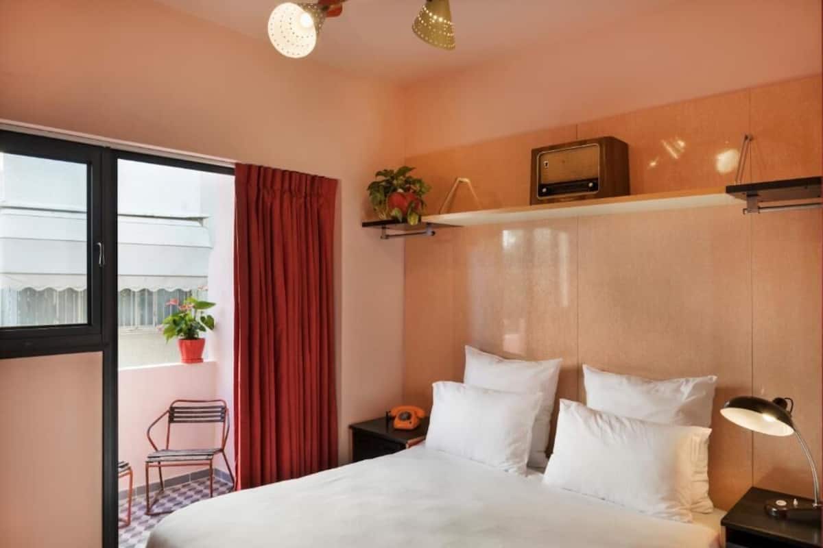 A hotel room with a bed, a nightstand, a radio, a potted plant, a chair, and a window with curtains.