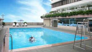 A swimming pool with a person swimming, lounge chairs, and a building with a glass facade. - Golden City Hotel (Foshan)