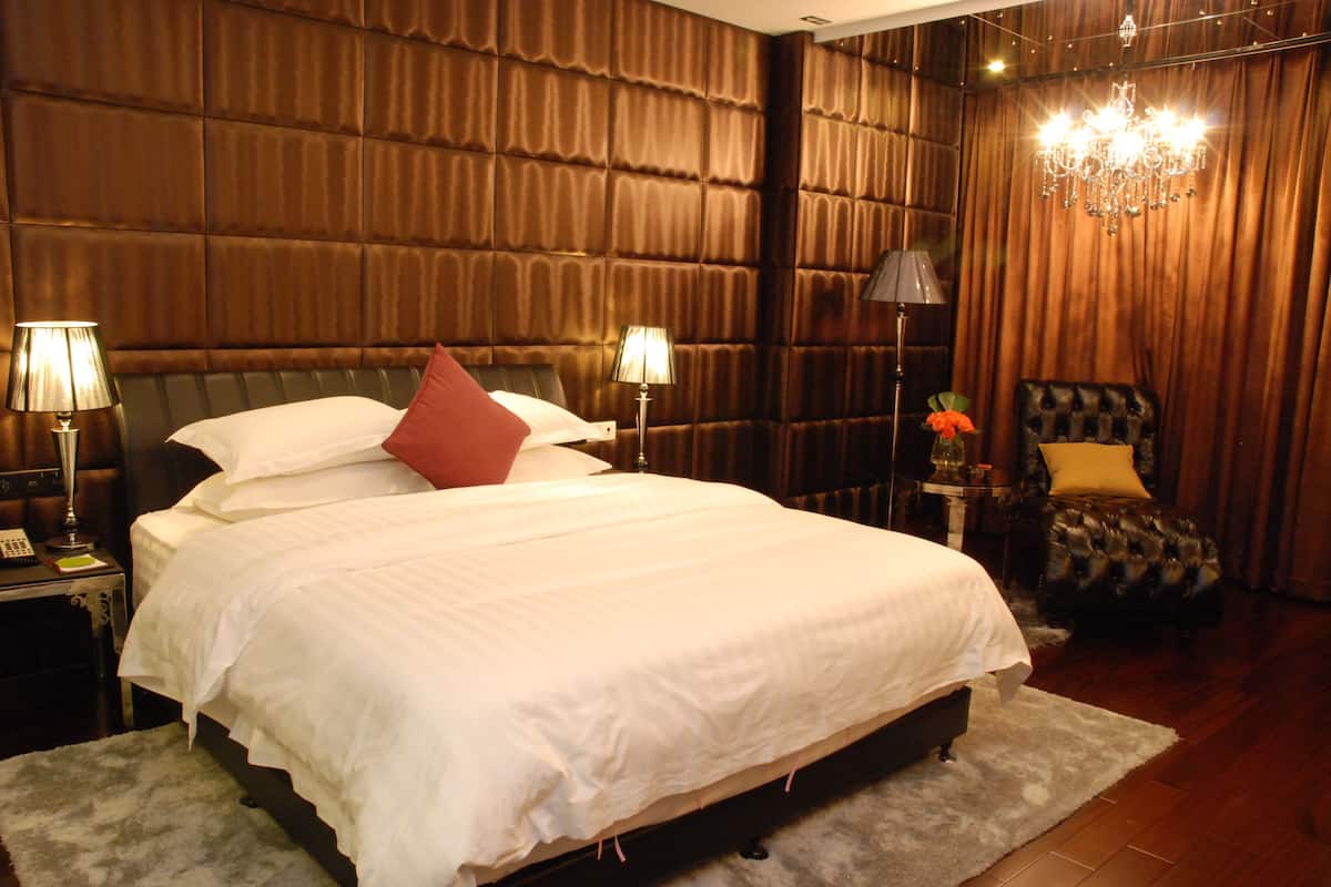 A hotel room with a large bed, two bedside lamps, a chandelier, a small table with a vase of flowers, and a leather armchair.