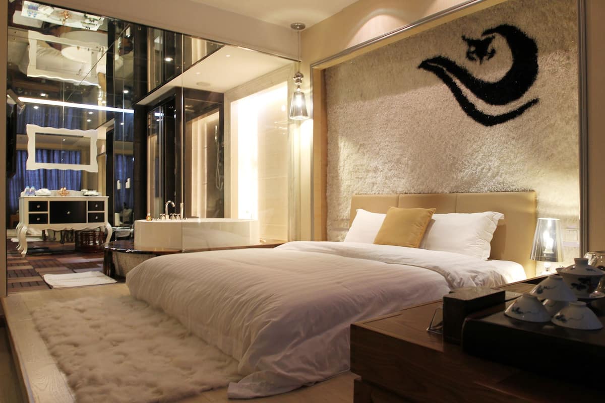 A modern hotel room with a large bed, a bedside table with a lamp, a mirror, and a decorative wall piece.