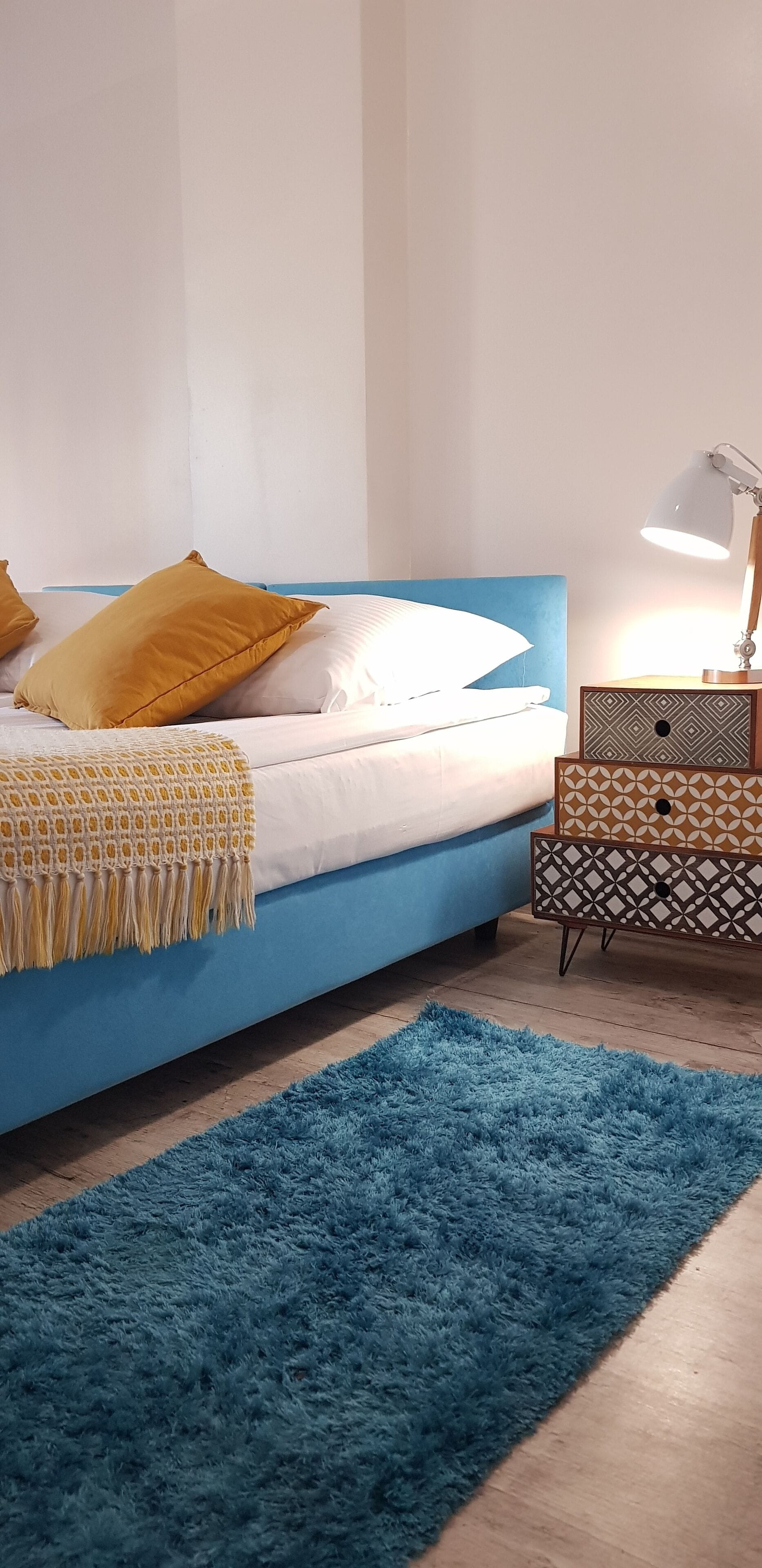 A bedroom with a blue bed, white and yellow pillows, a blue rug, and a bedside table with a lamp.