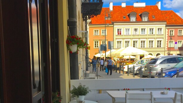 Un espace extérieur aménagé avec des tables et des chaises, un pot de fleurs et une vue sur une rue animée par des piétons et des voitures garées.