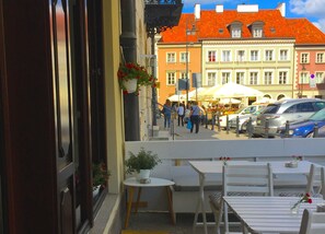 An outdoor seating area with tables and chairs, a potted plant, and a view of a street with pedestrians and parked cars. - Oki Doki OLD TOWN (Warsaw)
