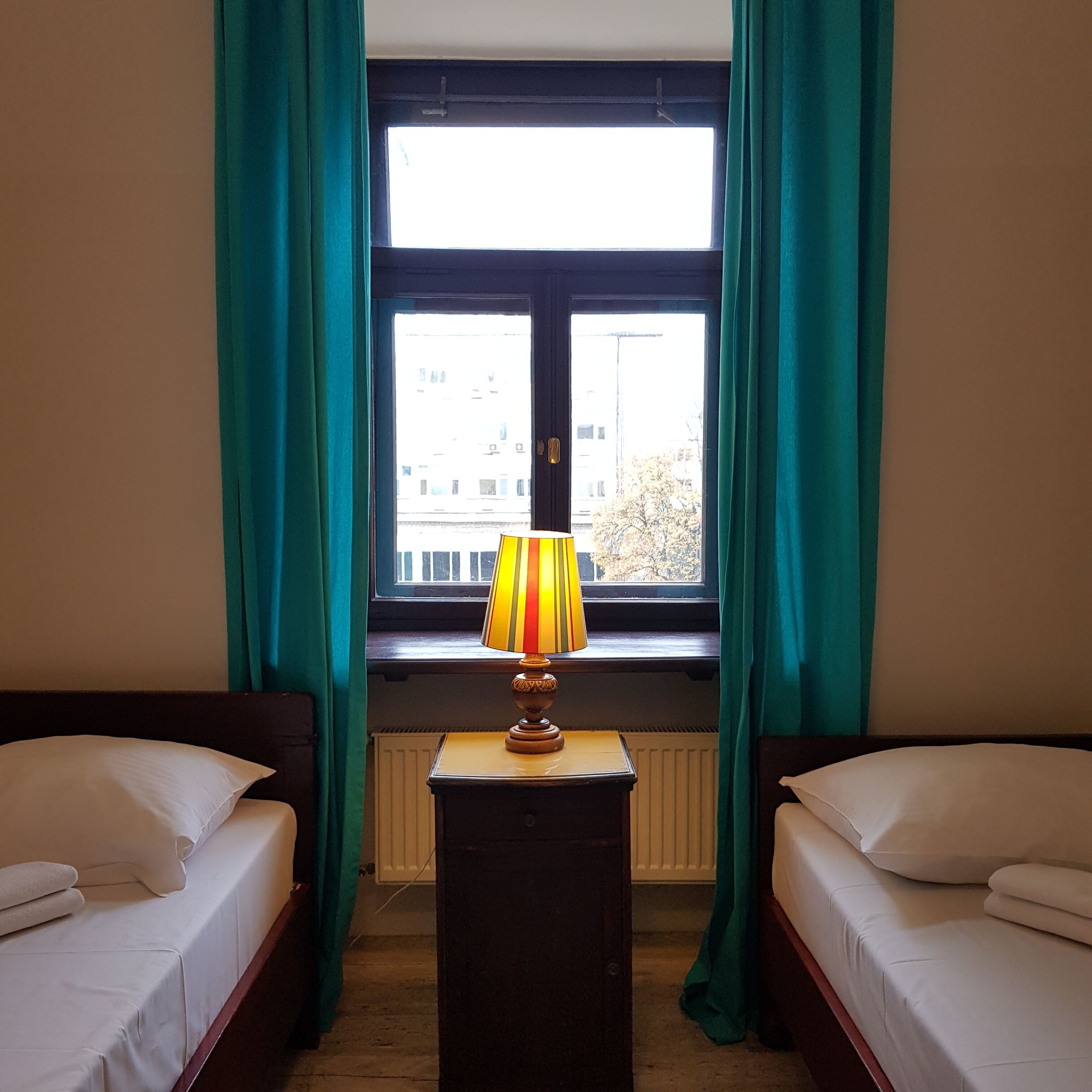 A room with two beds, a window with curtains, and a lamp on a table.