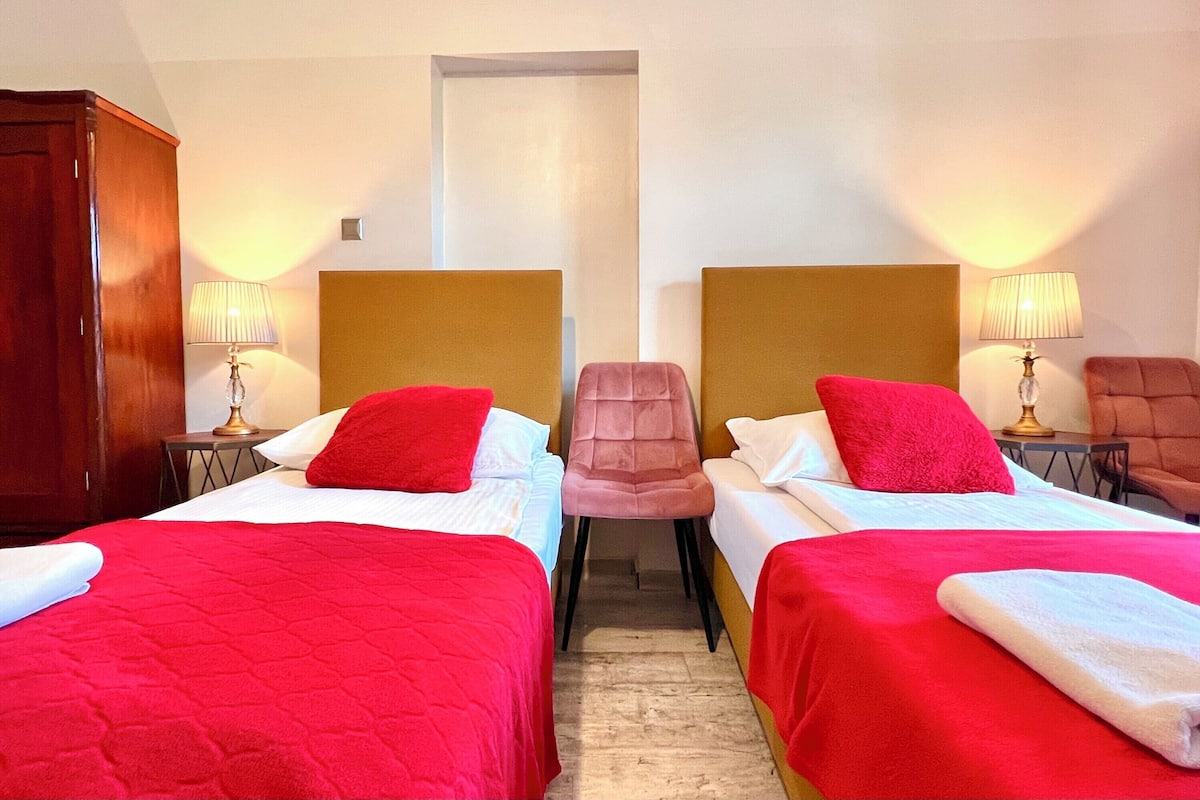 A hotel room with two beds, each with a red bedspread and white pillows. There are two lamps on either side of the beds, and a wooden wardrobe on the left.