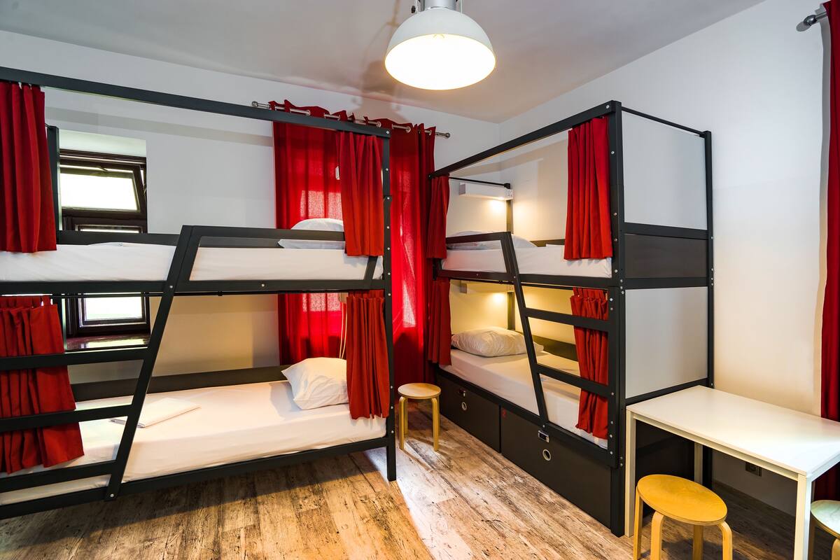 A bunk bed room with red curtains, a small stool, and a white table.